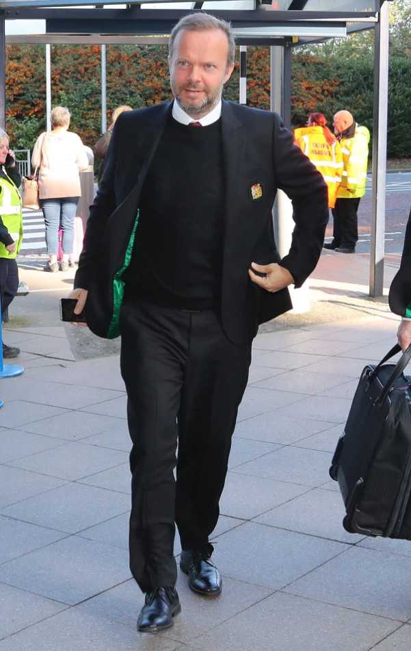 曼联CEO伍德沃德身着深色大衣，手提公文包，在贝尔格莱德机场步履匆匆，神情凝重，眼神中透露出对客场之旅的专注与思考。