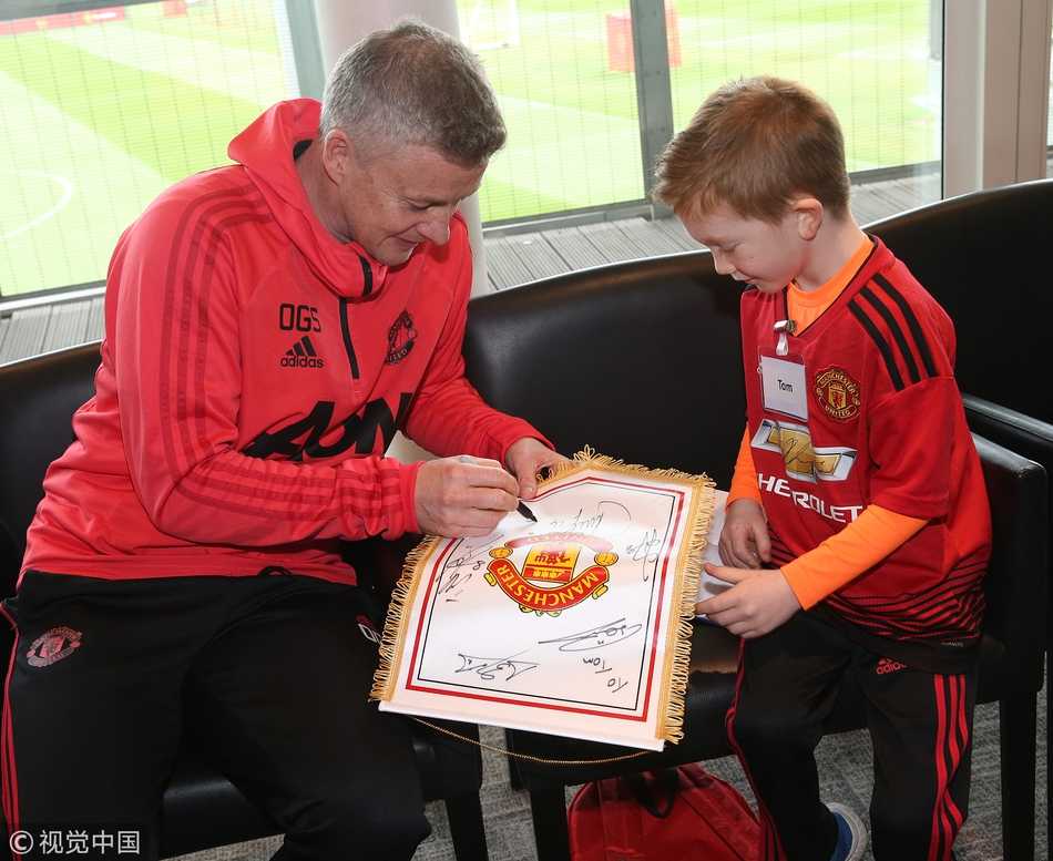 曼联球员马塔、斯莫林和林加德围成一个半圆，中间站着几个略显紧张的孩子。球员们正俯身倾听一个小女孩说话，女孩手里拿着一个曼联队徽，眼神里闪烁着激动的光芒，仿佛在讲述她与曼联的故事。