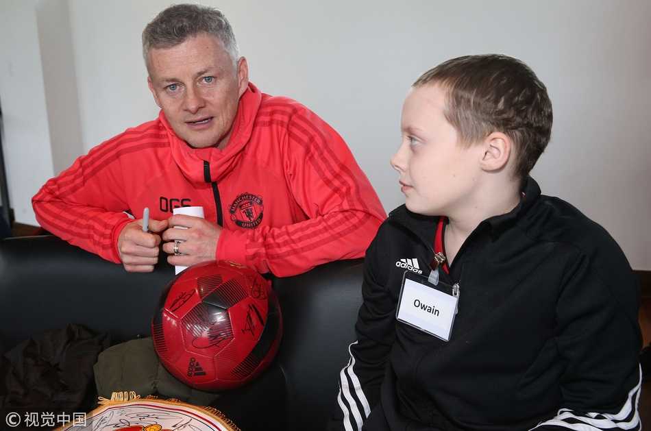 法国球星博格巴低头凝视着手中的一幅画像，那是他自己在球场上张开双臂庆祝的标志性动作。画作旁边，一位戴着眼镜的小男孩羞涩地笑着，博格巴伸出手臂搂着他的肩膀，画面定格在这一刻的惺惺相惜。