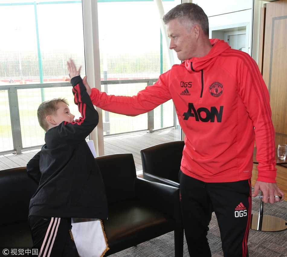 在一群身着曼联球衣的小球迷包围中，西班牙门神德赫亚露出了孩子般的笑容。他手里拿着一张由小球迷亲手绘制的画像，画中的他正做出标志性的扑救动作，旁边还有稚嫩笔迹写下的‘世界最佳门将’字样。