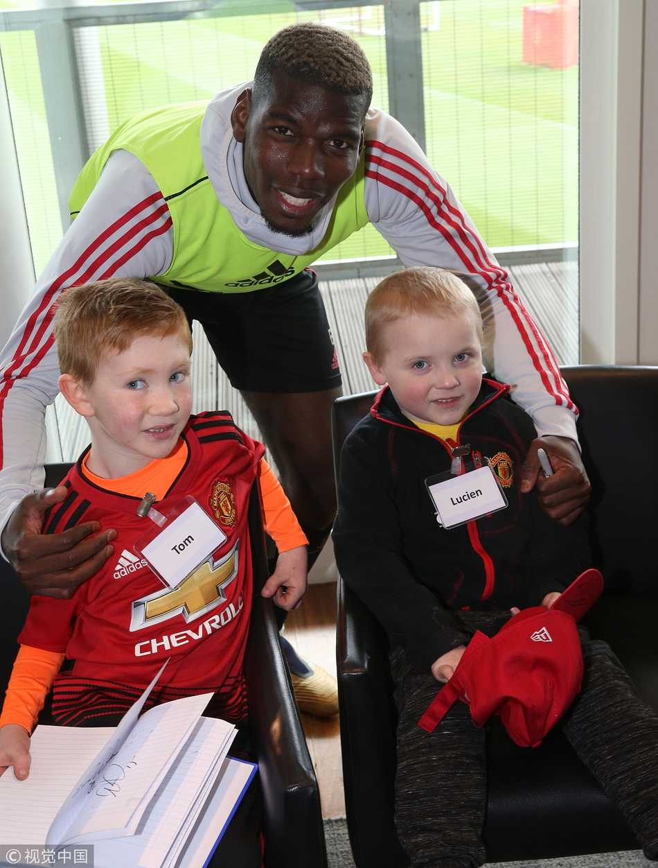 一位坐在轮椅上的小女孩穿着特制的曼联球衣，背号印着她的名字。她正举起手机，与刚刚签完名的曼联主帅索尔斯克亚合影。索尔斯克亚面带微笑，弯下腰配合小女孩的角度，背景是卡灵顿训练基地的绿茵场。