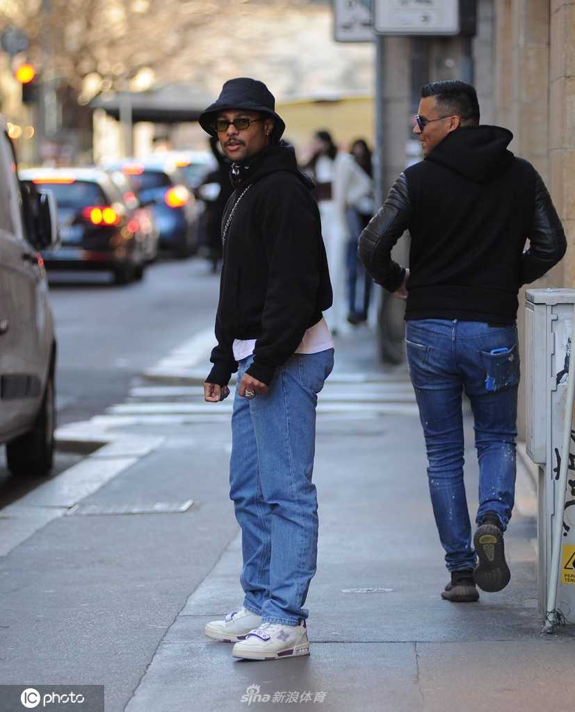 格纳布里侧身倾听友人交谈，阳光洒在肩头，其时尚的穿搭细节与米兰街景相得益彰