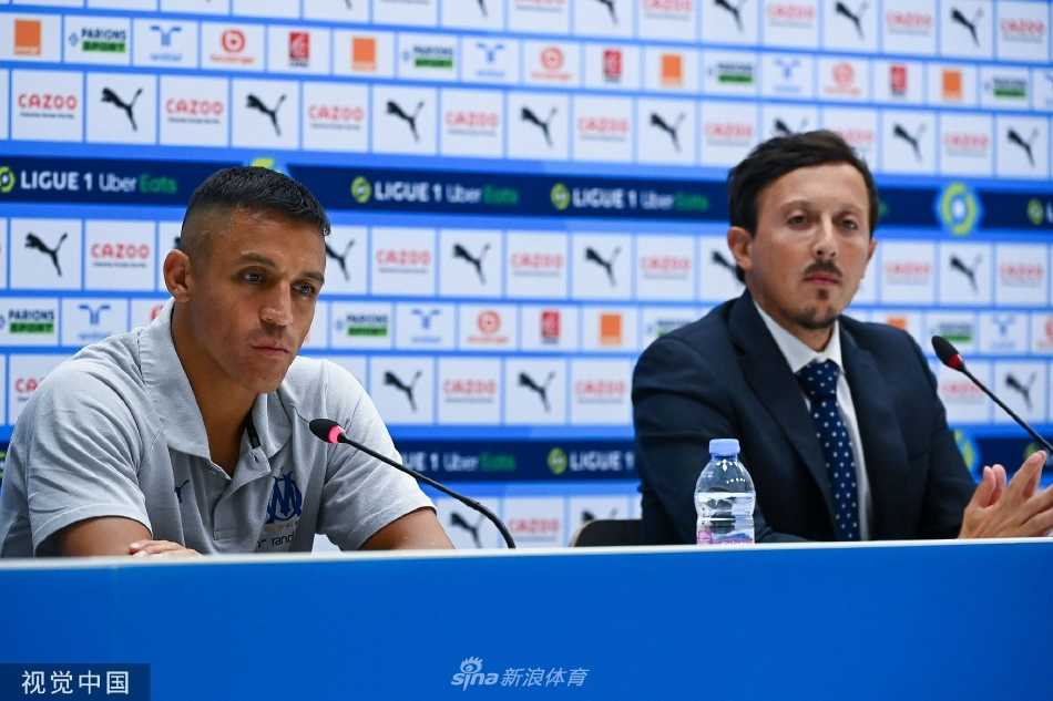 桑切斯与俱乐部官员一同站立，背景是马赛的主场球衣，他手指轻触下巴，似在深思未来的计划。