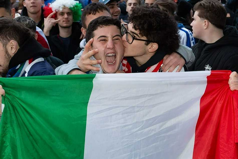 两位球迷勾肩搭背，脸上涂着代表意大利队的蓝色彩绘，眼神中闪烁着即将见证历史的期待与紧张，温布利大球场的灯光将他们身披的国旗映得格外鲜艳