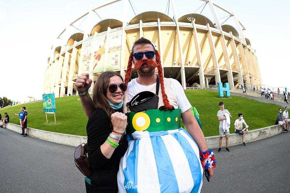 画面中的“粉红女郎”头戴鲜艳的花环，金色长发披肩，回眸一笑本想百媚生，但配上那略显沧桑的面容，却让周围的球迷笑得前仰后合，欢乐的氛围溢出屏幕。