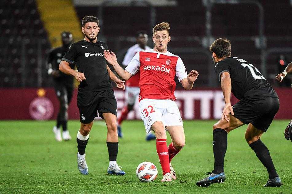 塞尔维特前锋格雷约翰·克耶伊在禁区内高高跃起，试图与兰斯后卫争顶头球，他面部肌肉紧绷，眼神中充满对进球的渴望，但在他身边，兰斯的防守球员如同铁塔般将他牢牢卡在身后