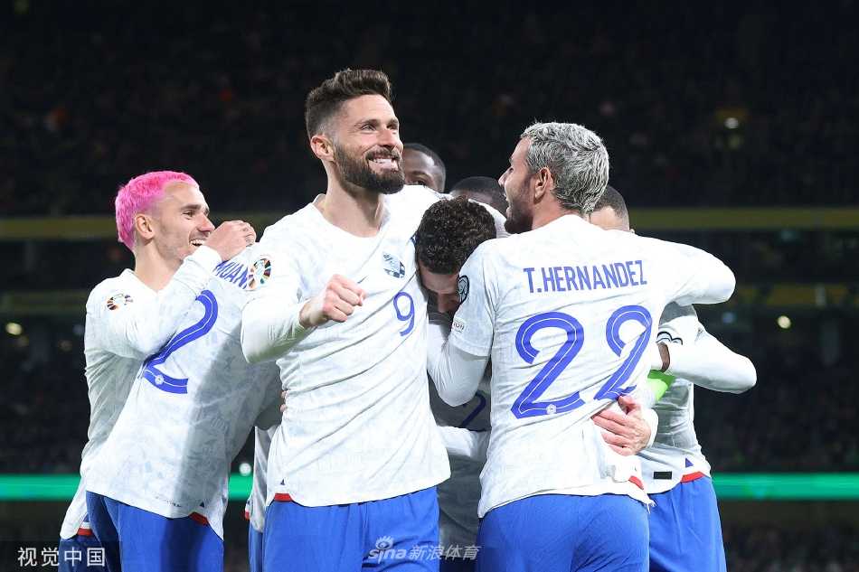 法国队全体队员围抱在一起庆祝进球，团队凝聚力在绿色的草地上绽放，主教练德尚在场边握拳呐喊。
