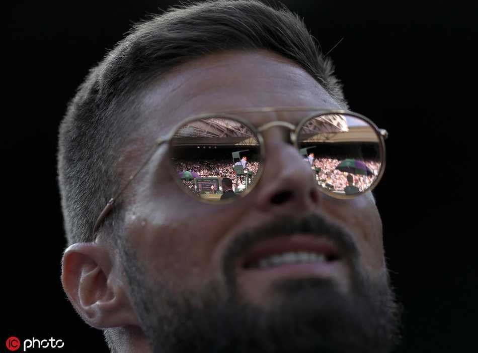 吉鲁在看台上露出标志性的憨厚笑容，双手准备鼓掌，身旁的观众也沉浸在温网决赛的紧张氛围中，这一刻他卸下锋霸的锐气，变身为一位享受比赛的普通球迷。