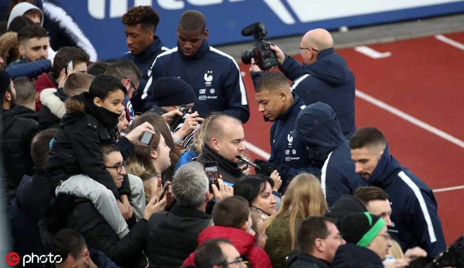 德尚教练在场边双手插兜，神情严峻地督战，远处是排队入场的球迷看台