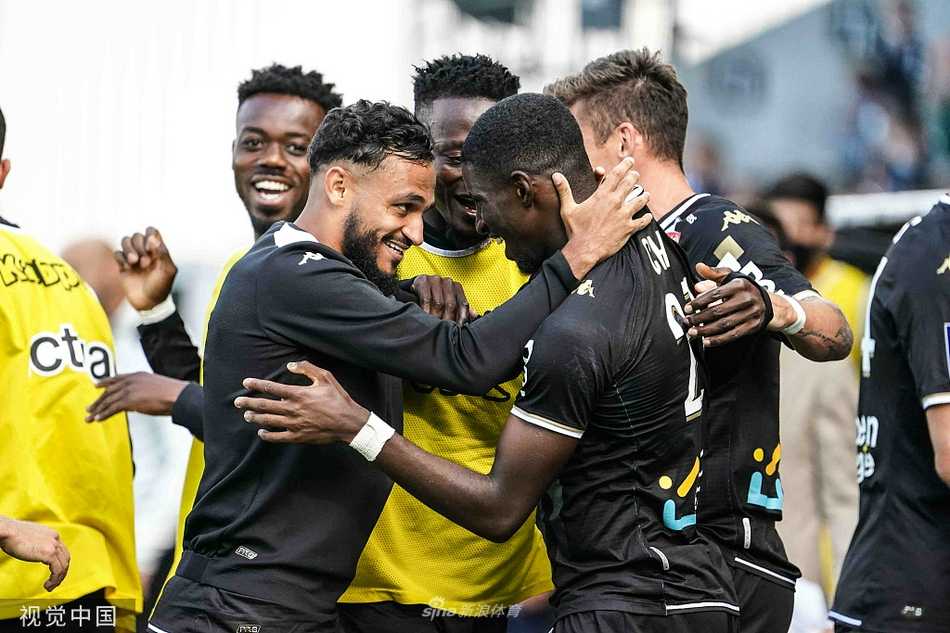 昂热球员阿里-乔破门后与队友在角旗区附近拥抱庆祝，看台上球迷们挥舞着旗帜和围巾，构成一幅胜利后狂欢的画卷