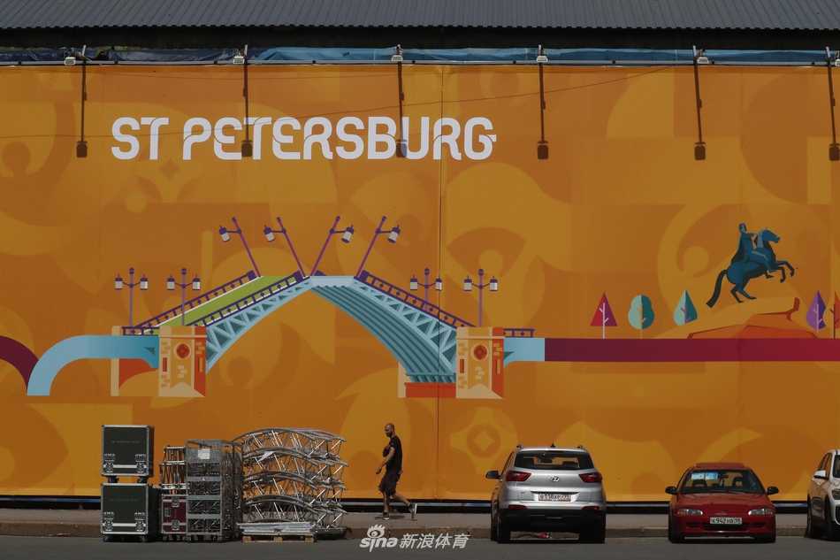 从高处俯瞰搭建中的圣彼得堡足球村，无数蓝色的施工围挡与未完成的白色帐篷模块规整排列，像是等待拼装的巨型积木，静静守望着不远处那座线条流畅的现代化球场