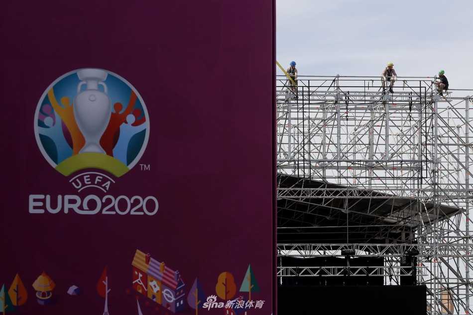 在夏日的阳光下，圣彼得堡足球村内，一名头戴安全帽的施工人员正专注地固定着最后一块围栏，他身后是已初具雏形的球迷嘉年华区域和远处若隐若现的泽尼特竞技场穹顶