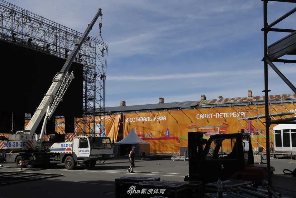 傍晚的夕阳为圣彼得堡足球村的施工现场镀上一层金色，一台高大的起重机矗立在画面中央，它的吊臂伸展在天空中，下方是忙碌的工人和排列整齐的临时建筑模块