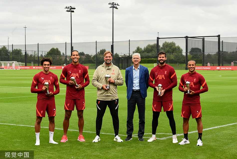 温格与克洛普在场边亲切交谈，两位战术大师面带微笑，气氛融洽，背后是利物浦训练基地的标语，预示着两代足球理念的友好碰撞。