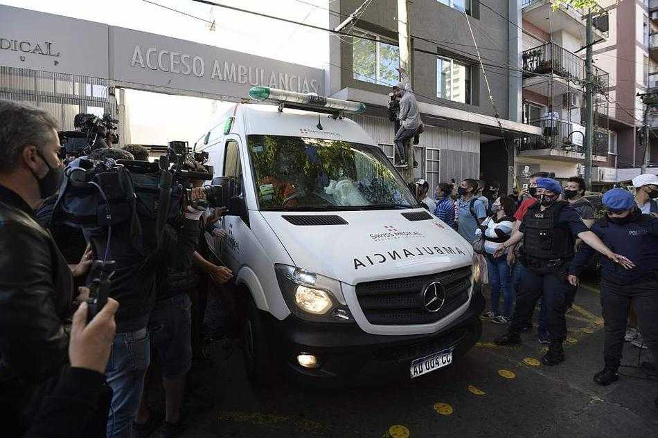 在一辆白色救护车的后门边，几名戴着口罩、穿着绿色医疗制服的工作人员神情紧绷，正将车内的情况与外界隔绝。车窗外，无数只手举着手机，试图穿透那扇紧闭的车门，捕捉到哪怕一丝球王的身影。人群的目光紧紧锁定在救护车深色的玻璃上，仿佛那里藏着整个阿根廷的希望。