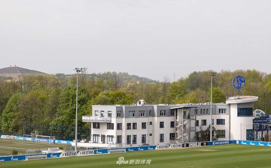 总部门口一侧，巨大的沙尔克04队徽下，疫情期间的临时指示牌显得格外醒目。蓝色的围栏如同屏障，将人们与这座充满历史感的建筑暂时隔离，唯有门前的绿植还在倔强地生长，象征着永不熄灭的矿工之魂。