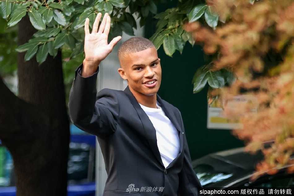 马利克·蒂奥身穿AC米兰经典红黑间条衫，手持印有自己名字的球衣，在米兰内洛训练基地的标志前展露自信而坚定的笑容。