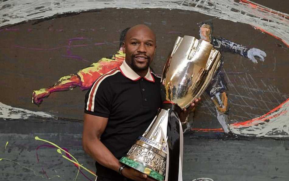 拳坛传奇梅威瑟身着一袭休闲华服现身都灵，嘴角挂着标志性的神秘微笑，右手比出招牌姿势，眼神中流露出对胜利者的惺惺相惜，身后尤文图斯的黑白剑条旗为他作衬。