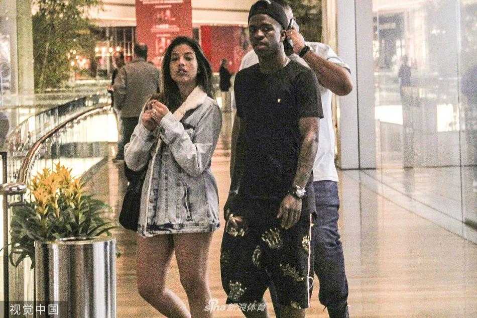 维尼修斯身穿简约的白色T恤和黑色短裤，头戴棒球帽，一手搂着女友的肩膀，两人在商场明亮宽敞的走廊里并肩前行，脸上带着轻松愉快的笑容，仿佛在讨论着接下来要去哪家店铺。