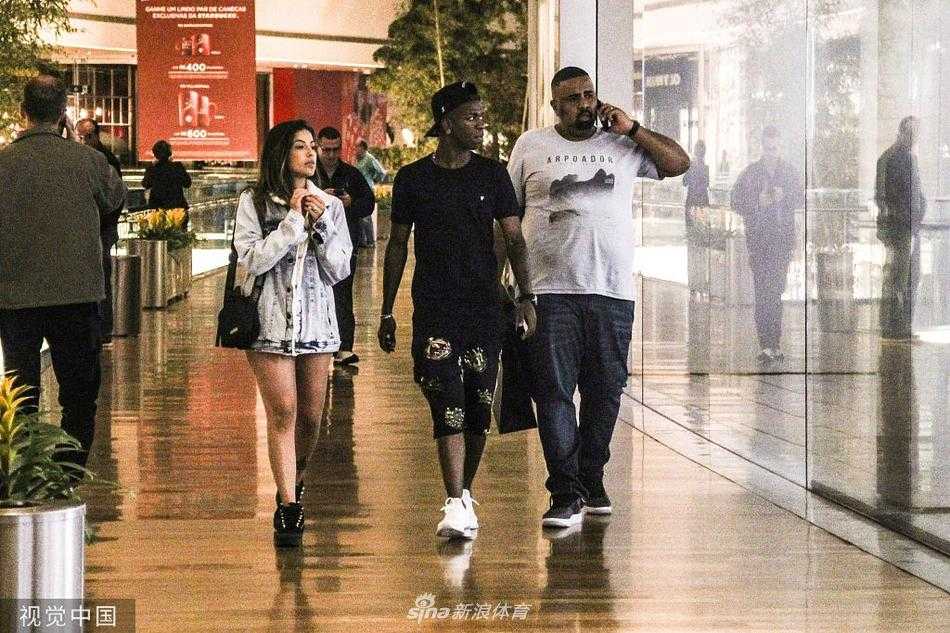 在一家装修精致的品牌专卖店柜台前，维尼修斯的女友正专注地俯身看着橱窗里的商品，似乎在比较着什么。维尼修斯则安静地站在她身后半步的位置，双手插在短裤口袋里，温柔地看着女友的侧脸，耐心十足地等待着她的决定。