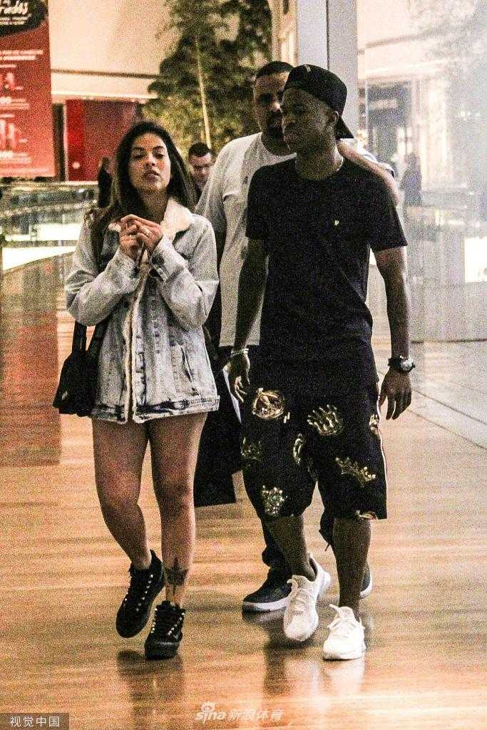 购物告一段落，维尼修斯和女友坐在商场内的休息长椅上。维尼修斯手里拿着几袋刚刚购买的物品，双腿随意地伸展着，神情放松。他的女友侧身坐着，两人似乎正在聊着什么有趣的话题，维尼修斯脸上露出了略带羞涩又开心的笑容，充满了少年感。