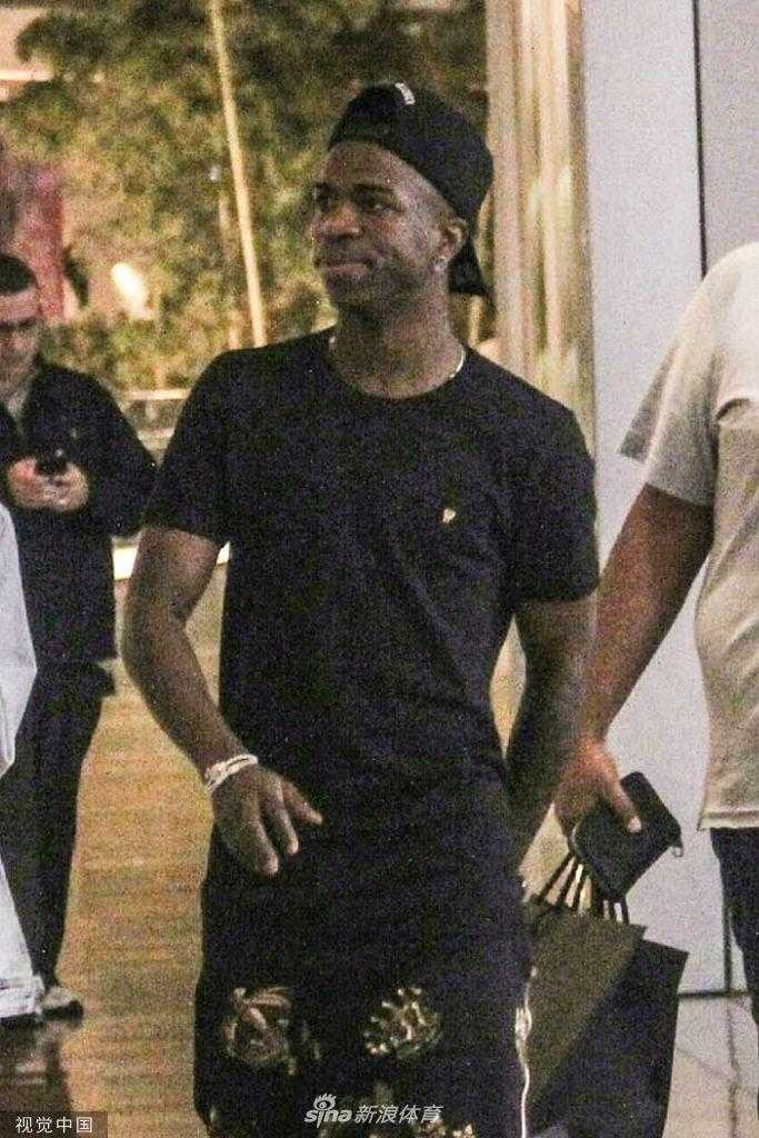 镜头拉近，维尼修斯的女友独自站在商场过道中，她身着一袭修身的米色连衣裙，搭配简约的凉鞋，展现出姣好的身材和时尚品味。她一只手轻轻拨弄着头发，目光看向别处，气质优雅从容，仿佛是时尚杂志中的一页街拍。