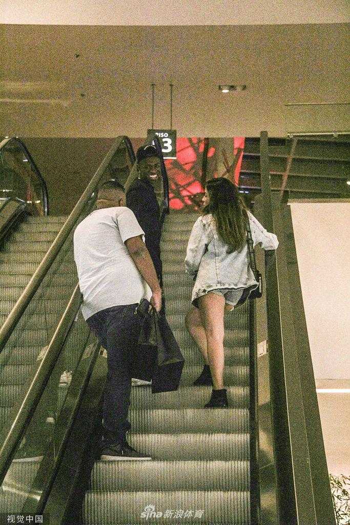 夕阳透过商场的玻璃穹顶洒下，维尼修斯与女友手牵手准备离开。他依旧戴着那顶标志性的棒球帽，女友的裙摆在行走中轻轻摆动。两人十指相扣，步伐一致，背影在温暖的余晖中拉长，为这次悠闲的约会画上了一个完美的句号。