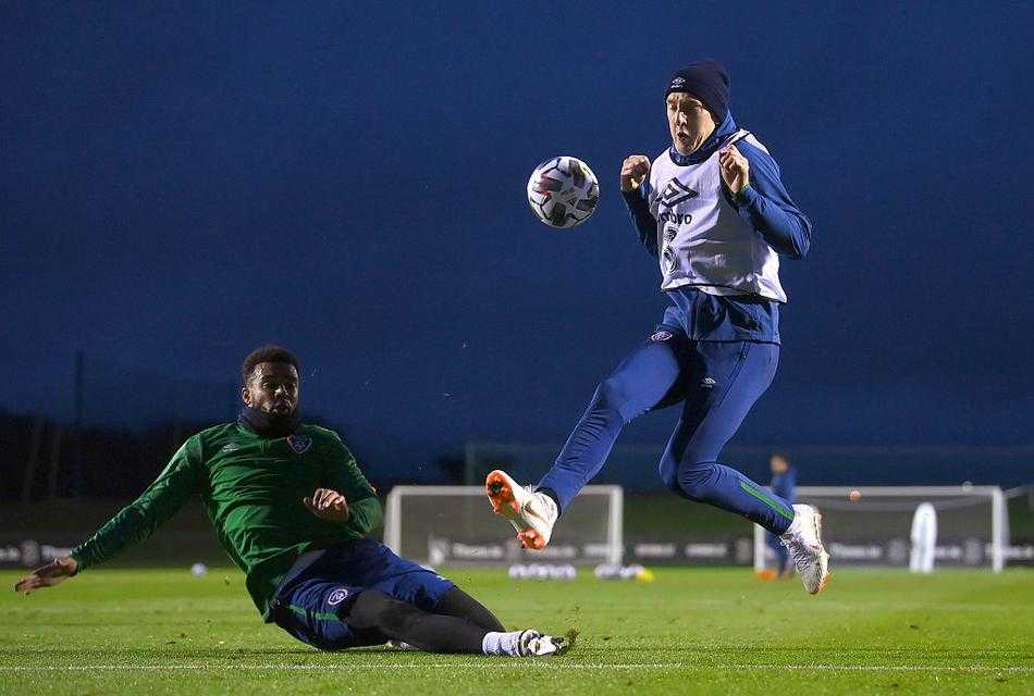 全景镜头俯瞰整个训练场，整支球队正在进行拉伸放松。绿色的草坪上，身穿荧光色背心的球员们排列得整整齐齐，场面颇为壮观。远处都柏林的城区天际线若隐若现，形成一幅动静结合的和谐画面。