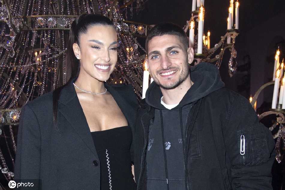 维拉蒂身着剪裁精致的深色大衣，内搭简约高领衫，爱妻杰西卡一袭优雅的奶油色长裙相伴，两人在时装周入口处驻足，维拉蒂侧身望向妻子，眼神里满是温柔与专注，仿佛周遭的喧嚣都与他们无关。