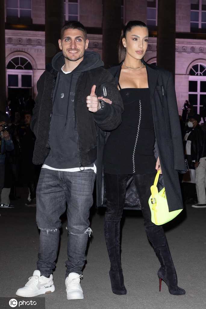 在时装周现场的斑驳光影中，夫妻二人并肩站立，维拉蒂的手轻轻搭在妻子背上，杰西卡手中握着手包，姿态放松而优雅，他们共同望向远方，仿佛在欣赏一场即将开始的秀，又像是在憧憬属于两人的未来。