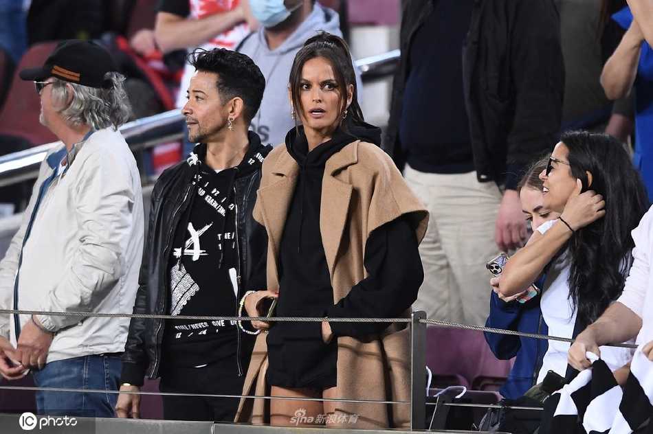 伊莎贝尔·古拉特身着简约黑色外套，手托下巴凝视球场，眼神中充满专注与期待，金色秀发在诺坎普的灯光下泛起温柔光泽。